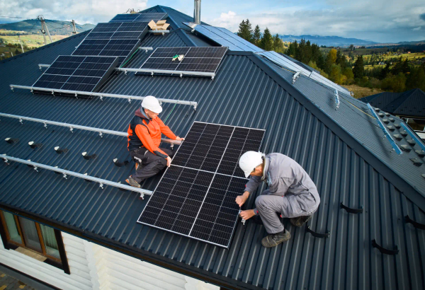 Zwei Handwerker montieren Solarmodule auf einem Hausdach mit Berglandschaft im Hintergrund